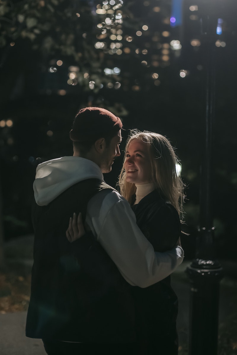 Side view of romantic couple cuddling looking at each other while standing on street against blurred trees and buildings at night time