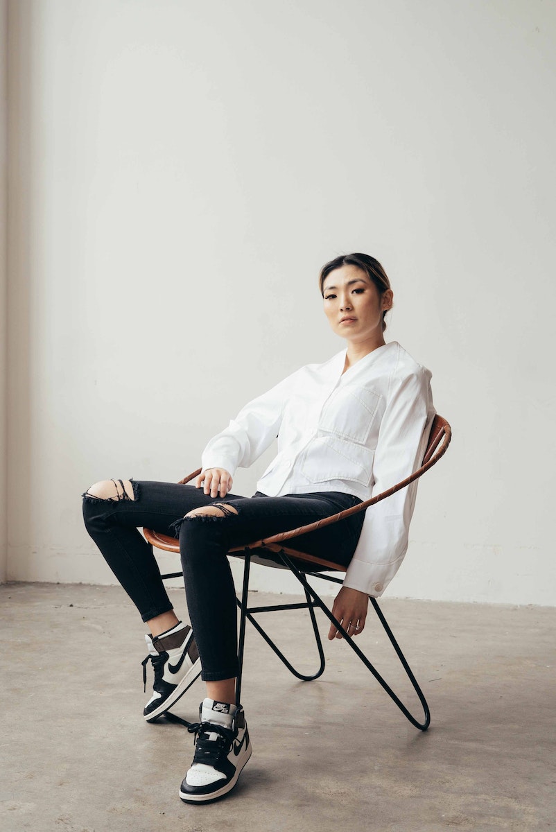Full body of confident ethnic female wearing ripped jeans with blouse and sneakers sitting in armchair in studio
