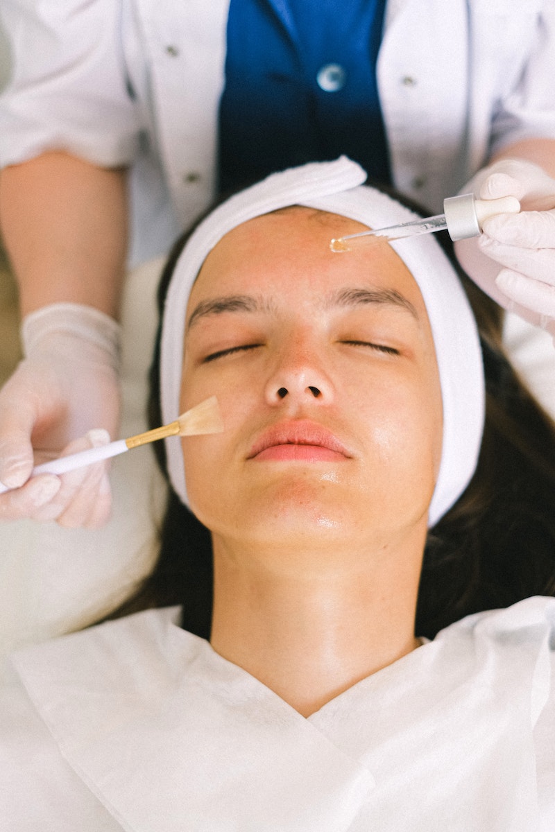 Top view skilled female cosmetologist in uniform and gloves applying essential oil on young woman face skin while working in contemporary spa center