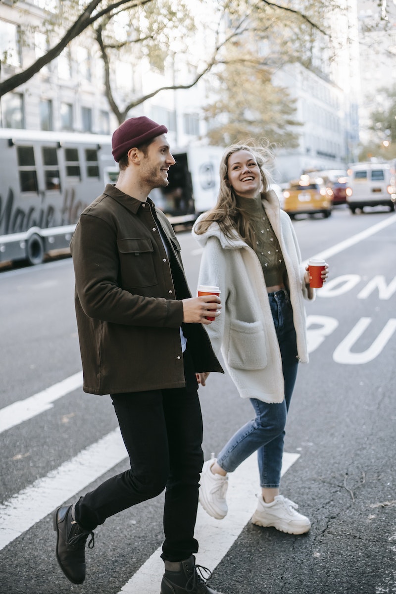 Young smiling couple in warm clothes strolling on city street with paper cups with drink and talking in daylight