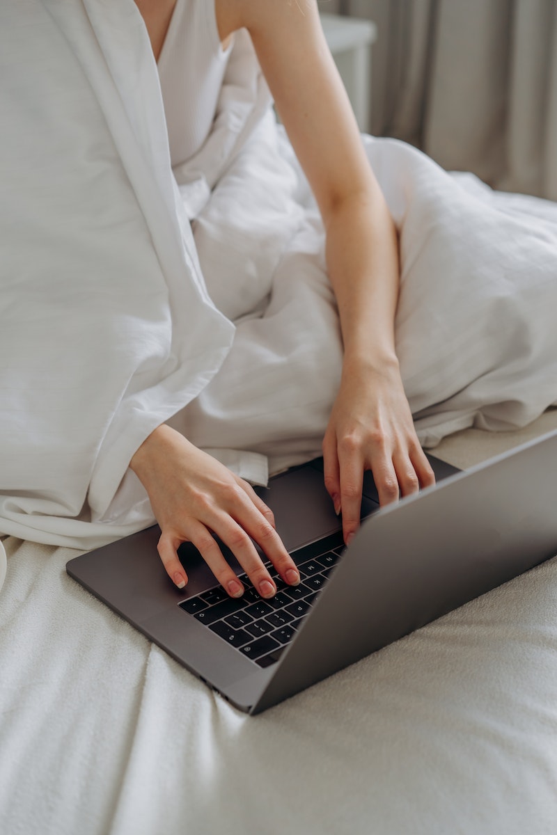 Person in White Sleeveless Top and Blanket Using Laptop on Bed