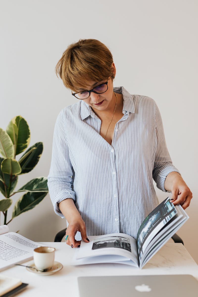 Serious female in casual light blue shirt standing near desk with laptop and cup of aromatic coffee while reading interesting magazine against white wall and houseplant