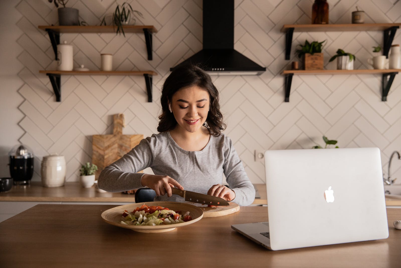 Cheerful ethnic female cutting fresh vegetables on cutting board while sitting at wooden table in kitchen with open portable computer in apartment