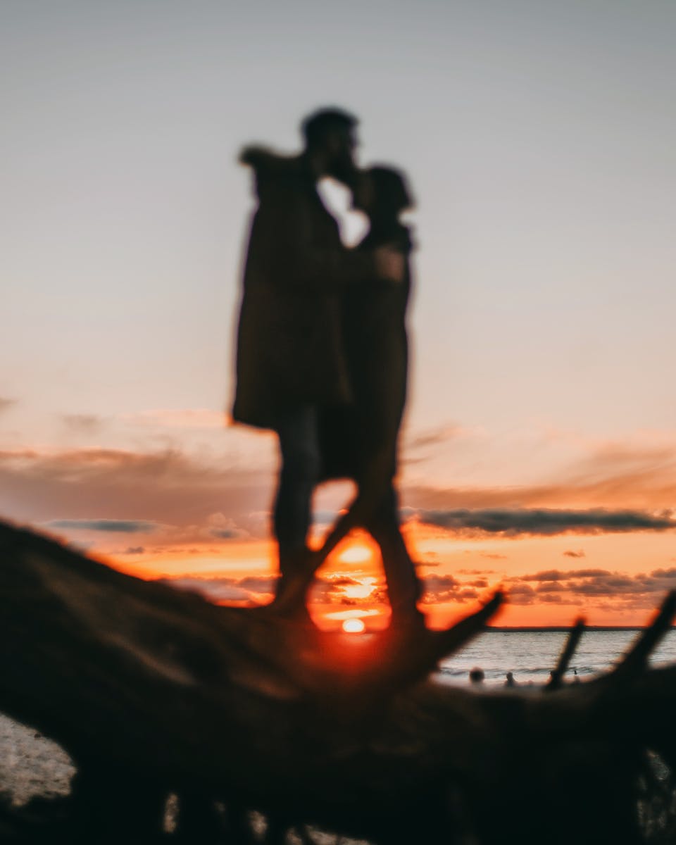 Defocused man cuddling and kissing unrecognizable woman in casual clothing while standing on coast of ocean against vibrant sky covered with orange clouds