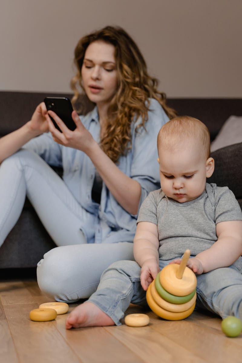 A Woman Using Her Mobile Phone while Sitting on the Floor Beside Her Baby