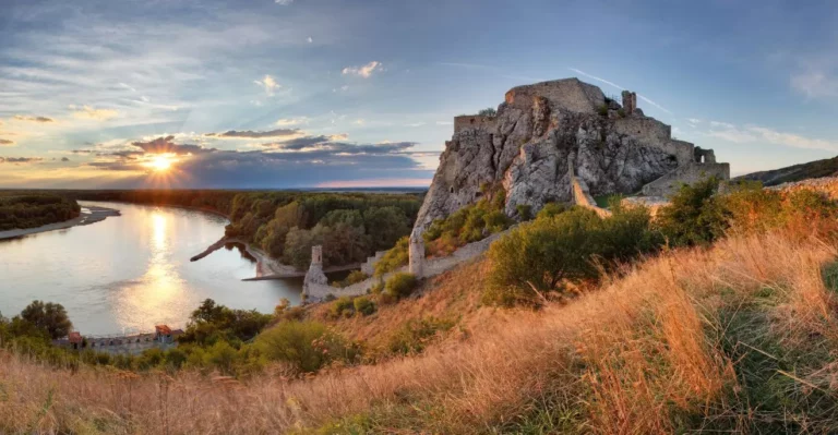 Hrad Devín pri západe slnka, Slovensko, rieka Dunaj a historické ruiny so zeleňou v pozadí.