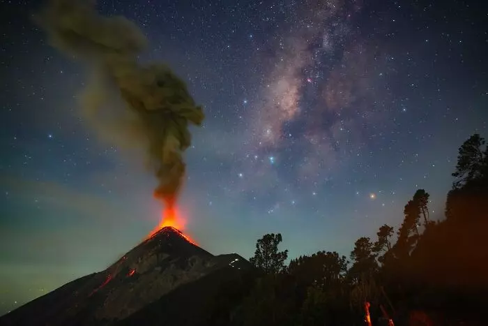 Erupcia sopky pod hviezdnou oblohou, vychádzajúci dym a láva osvetľujú nočnú krajinu s Mliečnou cestou.