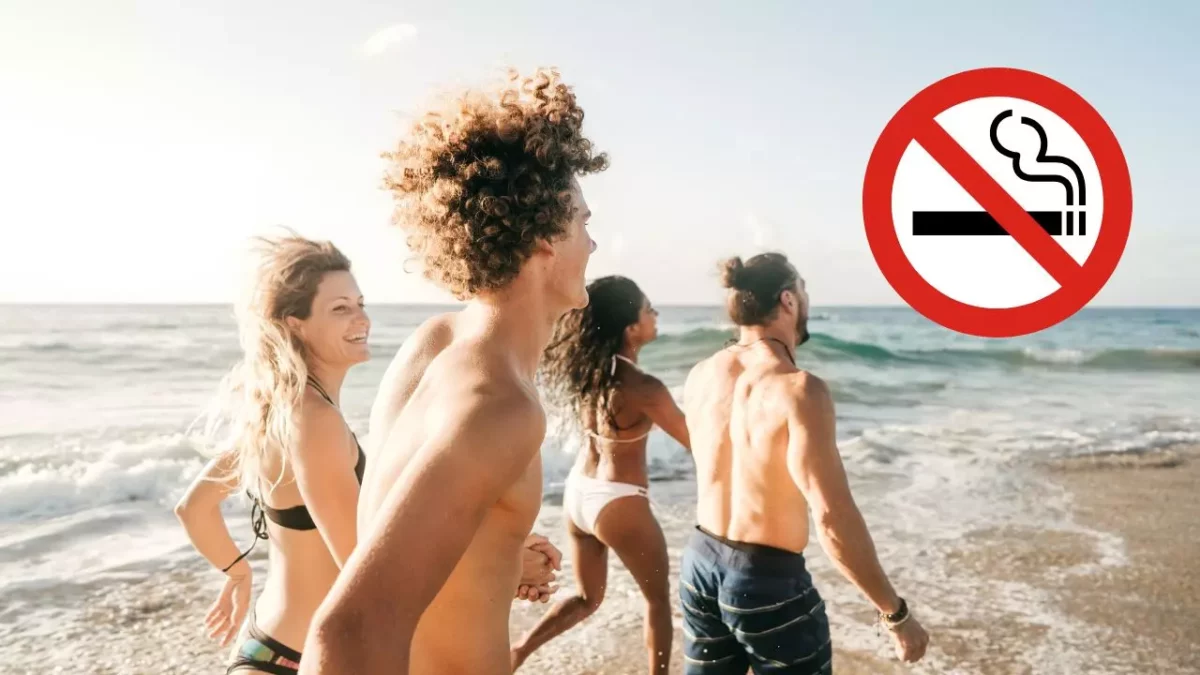 Skupina ľudí na pláži za slnečného dňa, symbol zákazu fajčenia v pravom hornom rohu.