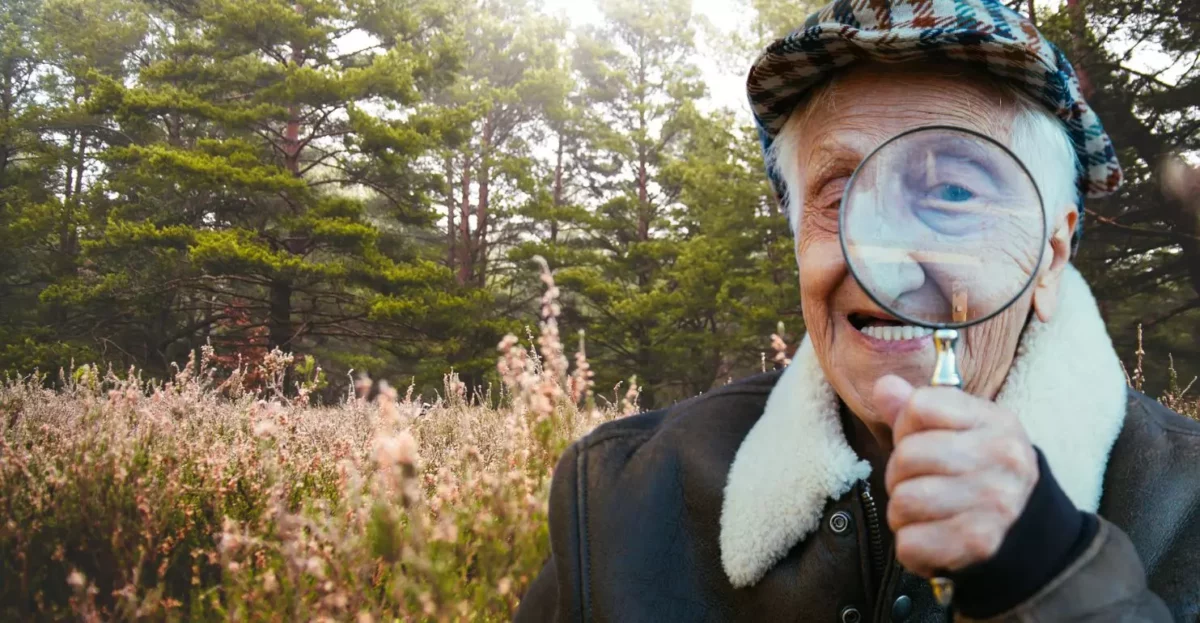 Starší muž s lupou v lese, usmievajúci sa počas skúmania okolia.