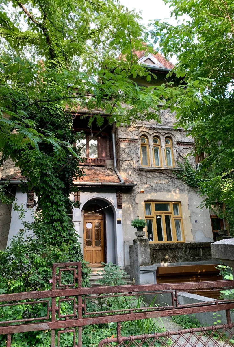 Starý dom obklopený zeleňou a popínavými rastlinami, s drevenými dverami a ozdobnými oknami.
