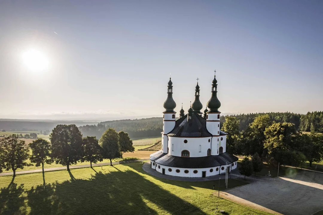 Kostol v prírode pri východe slnka, obklopený stromami a zelenou krajinou, s modrou oblohou v pozadí.