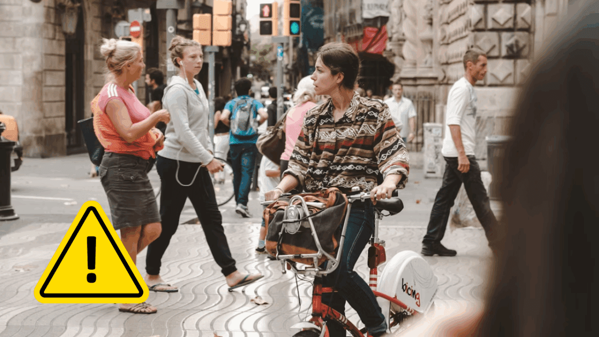 Žena na bicykli v rušnom mestskom prostredí, obklopená chodcami a dopravnými značkami.