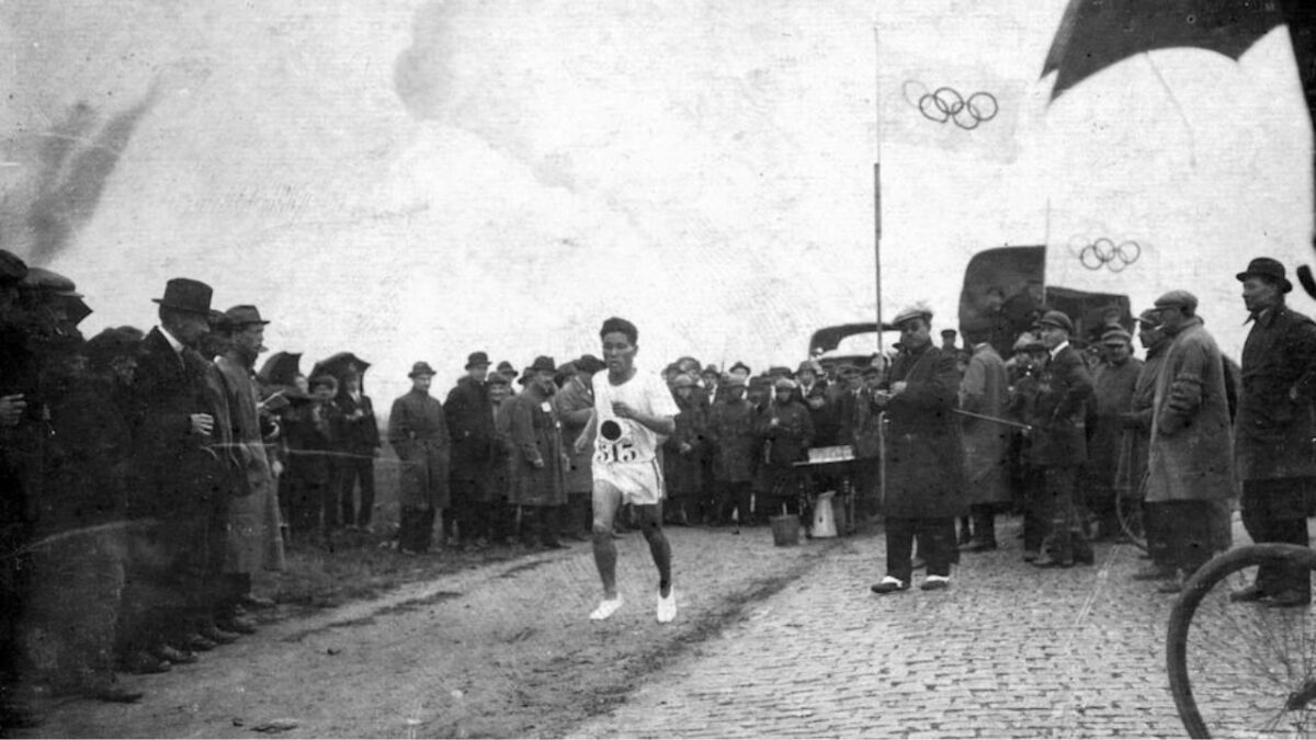 Bežec na olympijských hrách, starší čiernobiely záber, dav sleduje športovca na kamennej ceste s vlajkami v pozadí.
