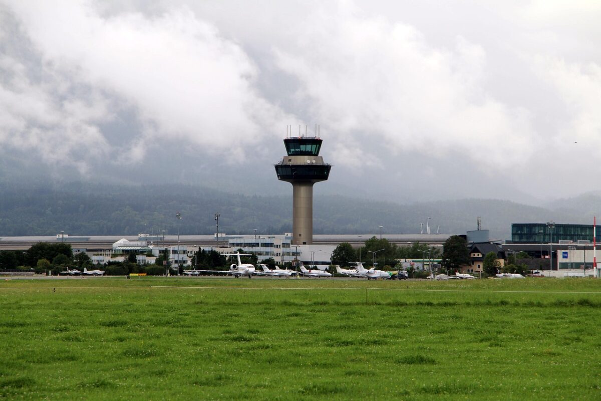 Letisková riadiaca veža obklopená prúdovými lietadlami na zelenom letisku v zamračený deň.