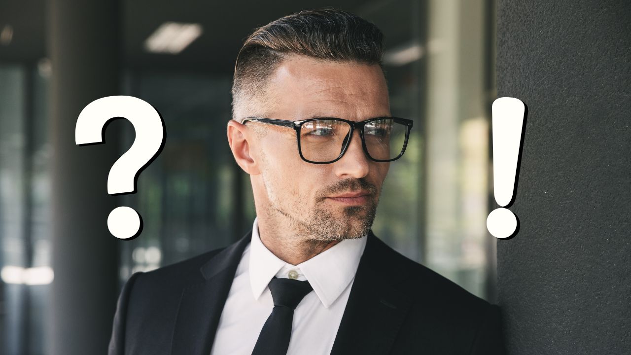 Biznis muž s okuliarmi pri stene v obleku s otáznikom a výkričníkom na pozadí.