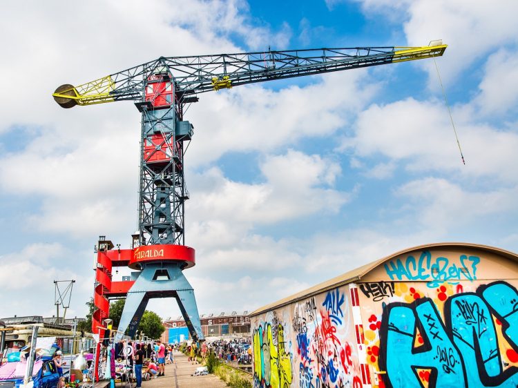 Žeriav na pozadí graffiti steny počas slnečného dňa na industriálnej ploche plnej ľudí.