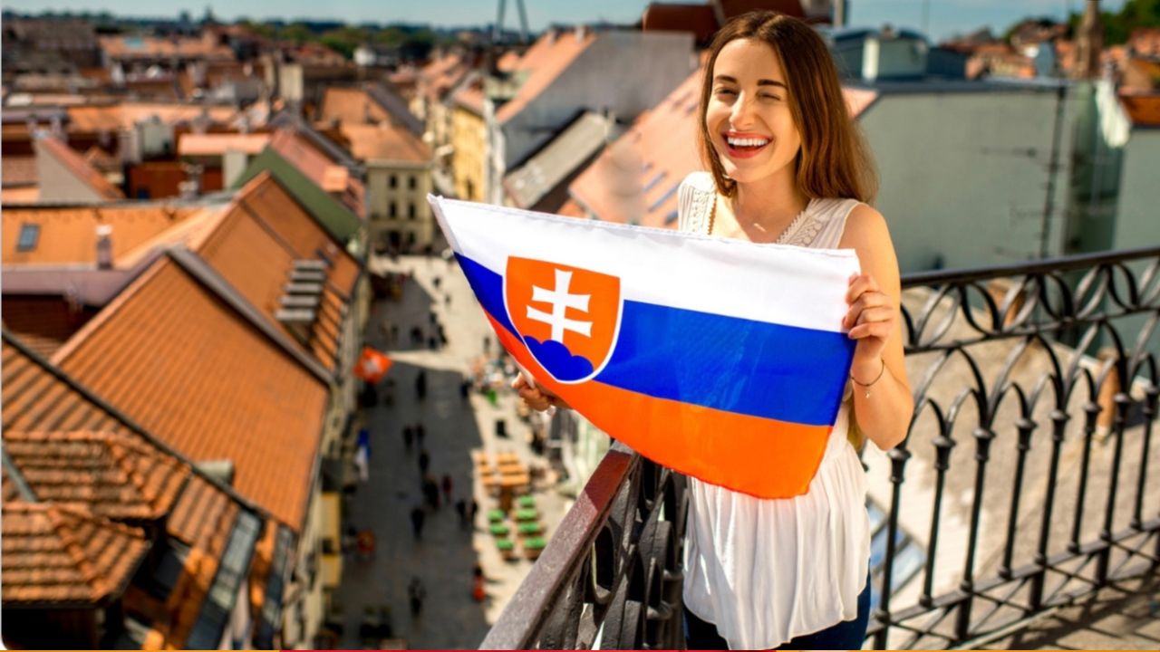 Žena drží slovenskú vlajku na historickom balkóne s výhľadom na mesto, letný deň.