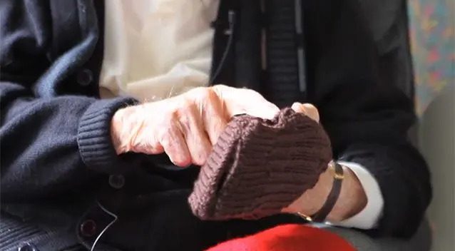 Staršia osoba pletie tmavohnedú čiapku, sedí v pohodlnom kresle oblečená v svetri.