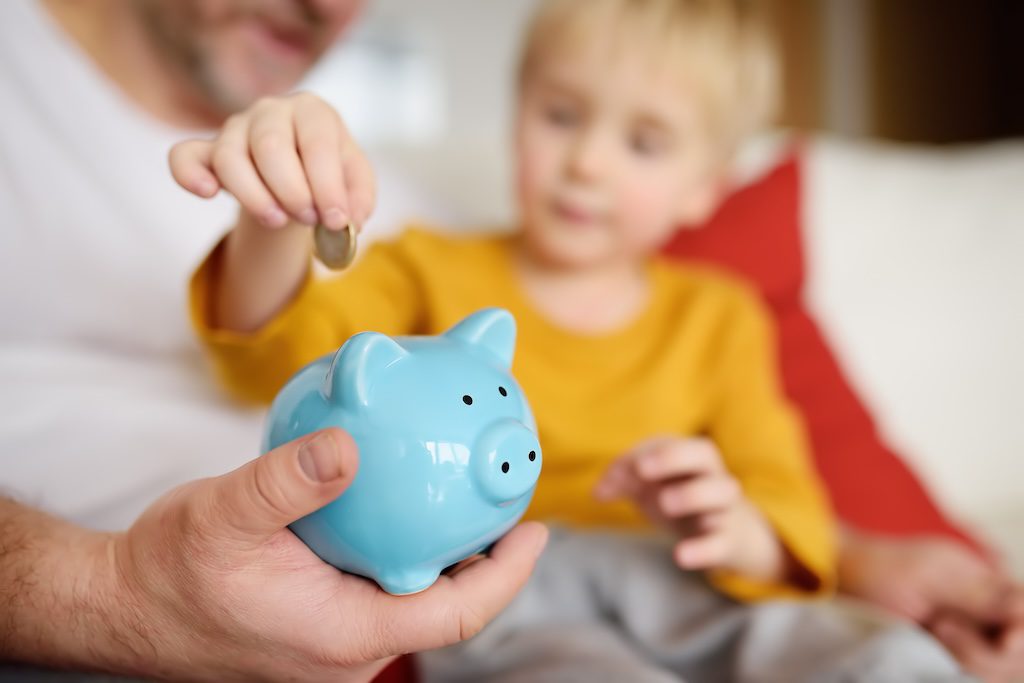 Dieťa vkladá mincu do modrej prasiatka ako symbol učenia sa sporenia a finančnej gramotnosti.