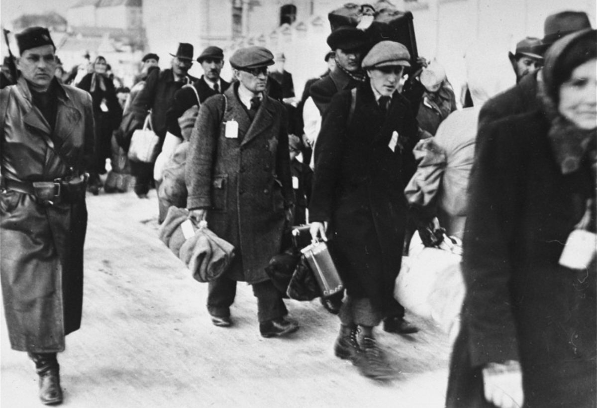Ľudia nesúci batožinu v zime, zachytení v čiernobielej fotografii historického kontextu.