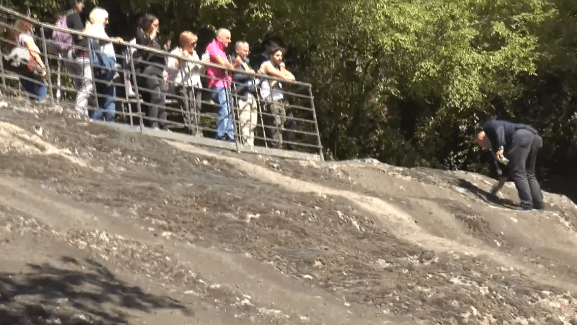 Skupina ľudí pozoruje muža s lopatou na svahu, obklopení zeleňou v prírodnom prostredí.
