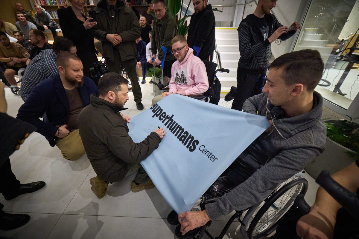 Skupina ľudí sa zhromažďuje okolo človeka podpisujúceho vlajku s názvom superhumans center.