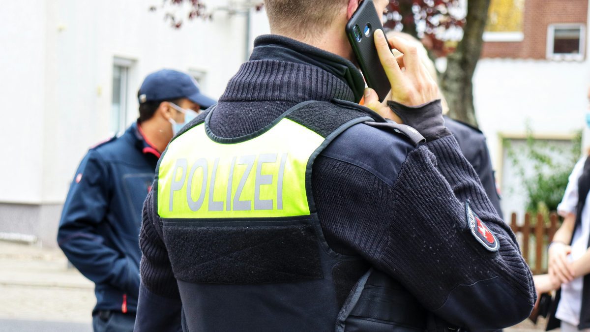 Policajt v uniforme telefonuje, okolo sú ďalší ľudia v exteriéri.