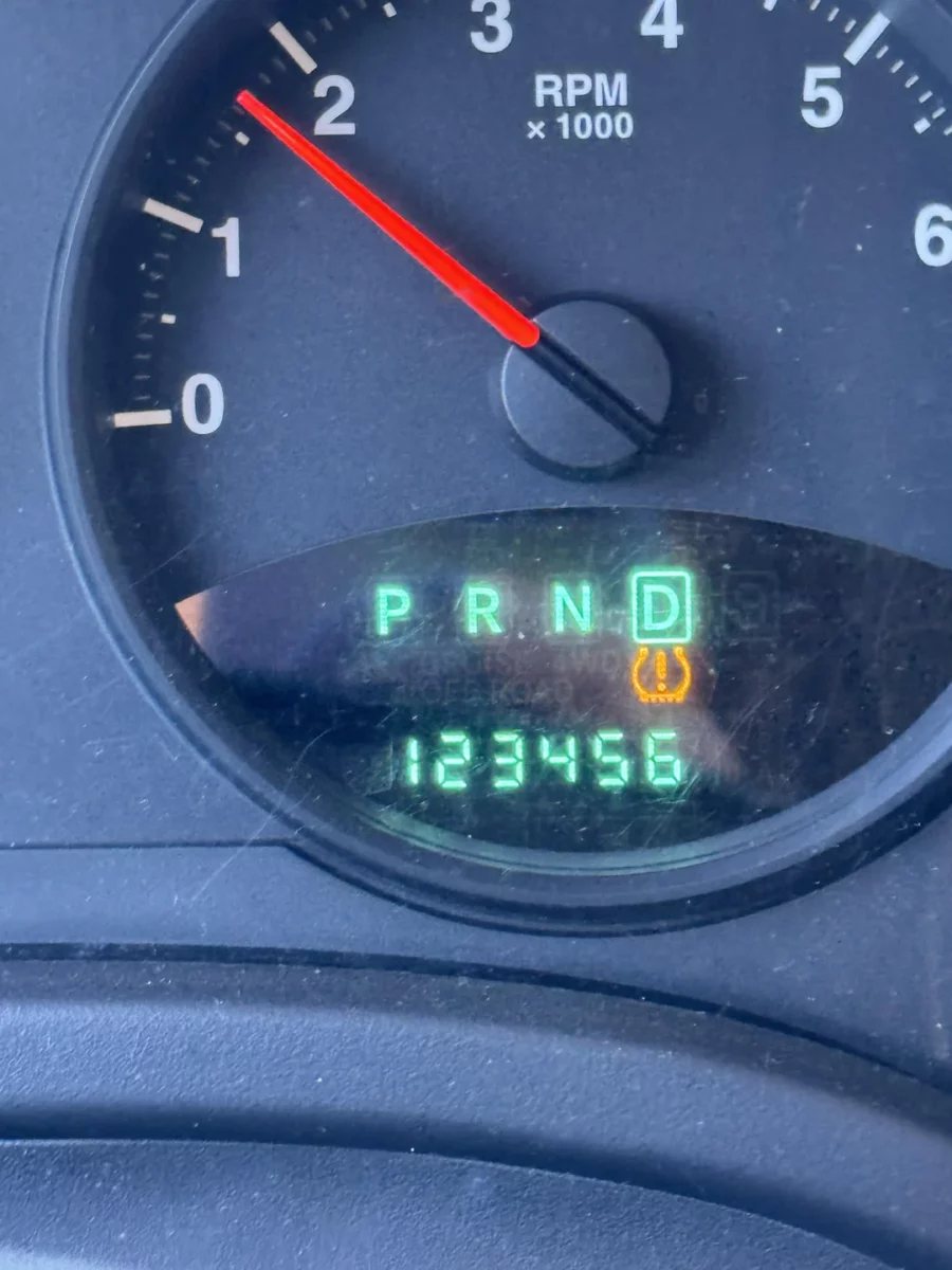 Automobilový tachometer ukazujúci 123456 km a symbol kontroly tlaku v pneumatikách.