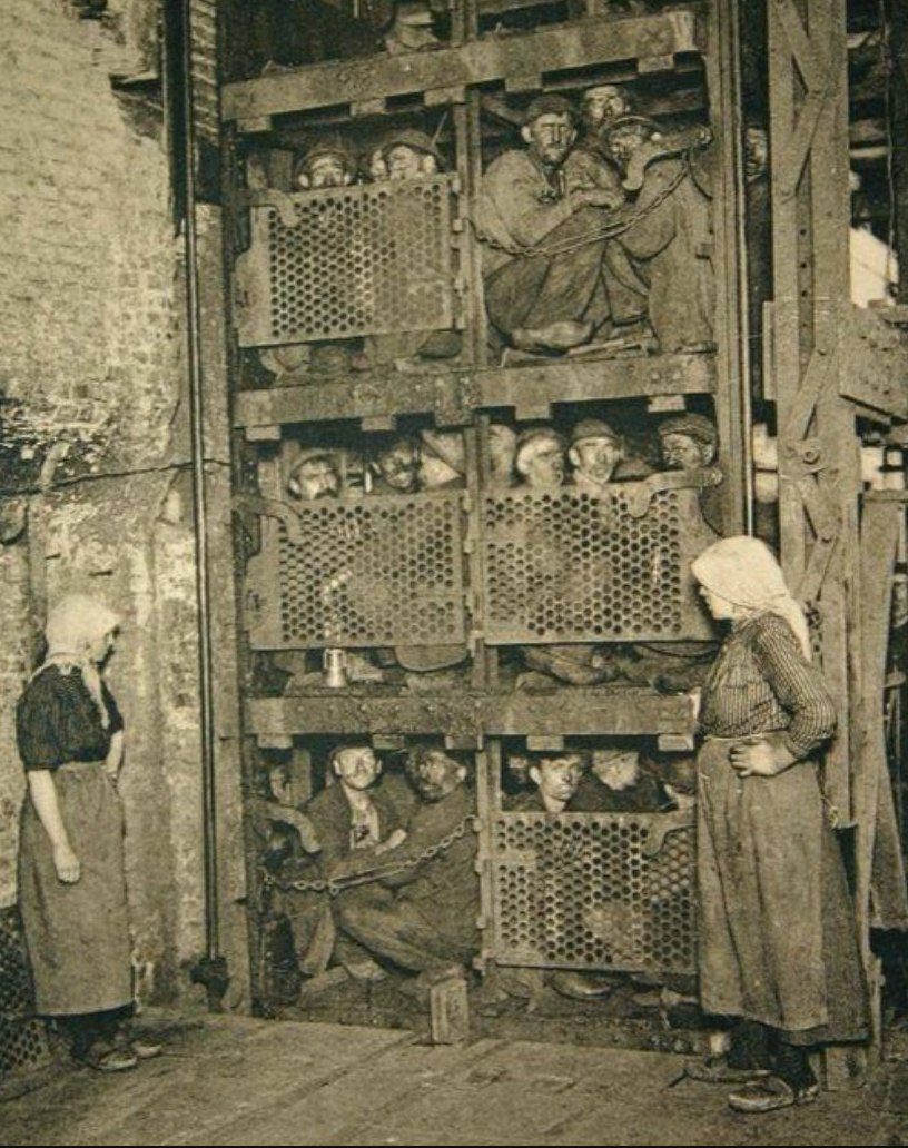 Banskí robotníci v preplnenej klietke čakajú na prepravu; historický pohľad na pracovné podmienky v baníctve.