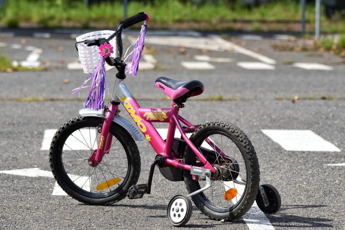 Ružový detský bicykel s pomocnými kolieskami na parkovisku, ozdoby na riadidlách.