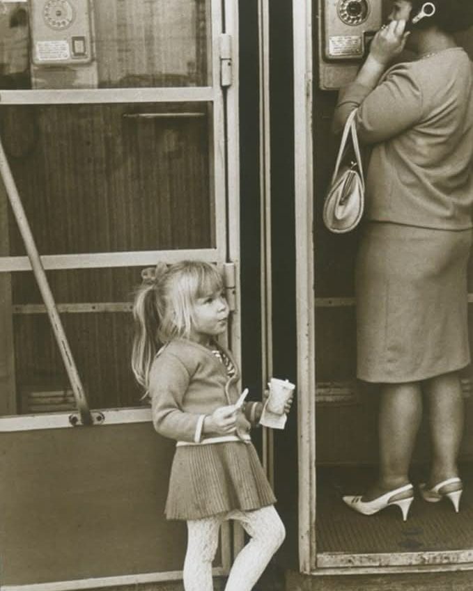 Dievčatko čaká pri telefonnej búdke, zatiaľ čo žena telefonuje. Retro atmosféra a detská zvedavosť.