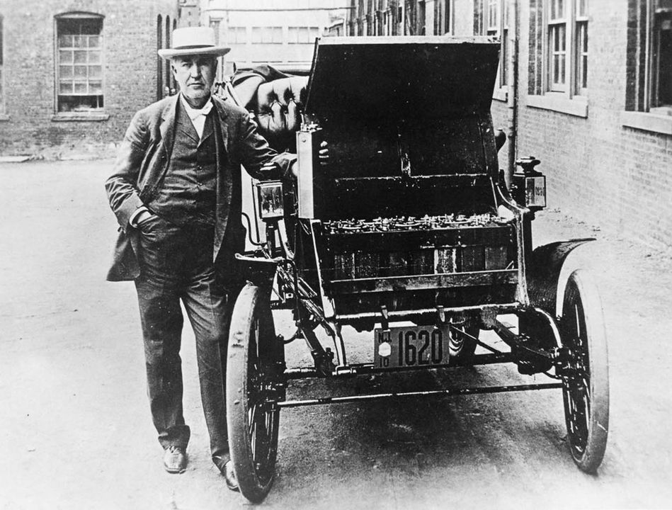 Muž stojí vedľa historického automobilu na dláždenej ulici, oblečený v obleku a klobúku.