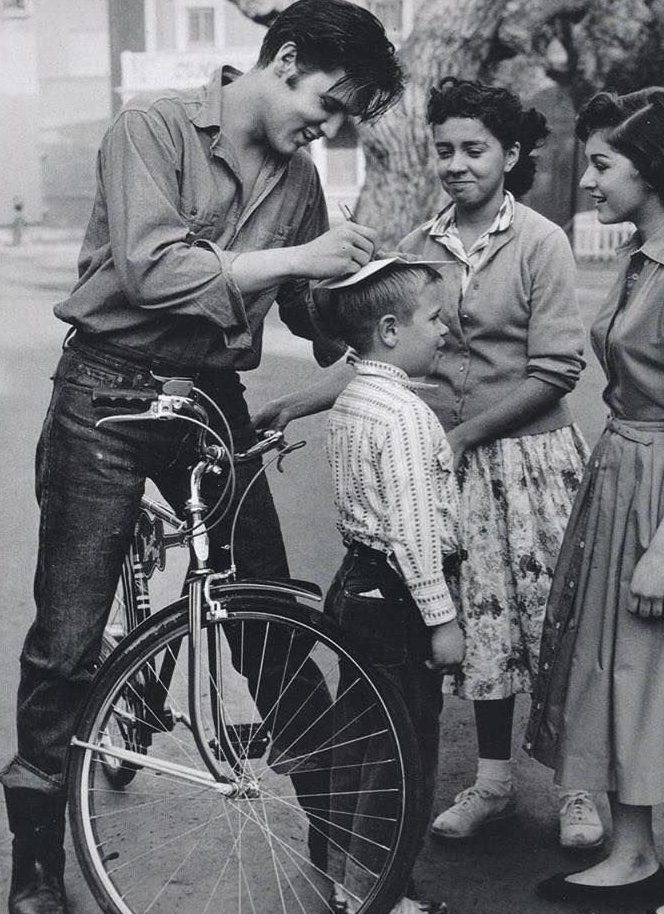 Muž podpisuje klobúk chlapcovi, vedľa stojí bicykel a dve dievčatá sa usmievajú. Vintage pouličná scéna.