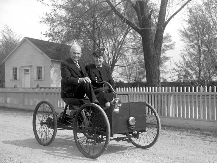 Starý automobil s dvoma osobami, v pozadí vidiecky dom a stromy, čiernobiela fotografia.
