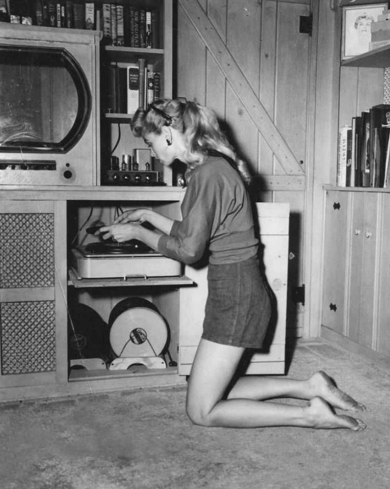 Žena na kolenách ovláda retro gramofón v vintage miestnosti so starými knihami a televízorom.