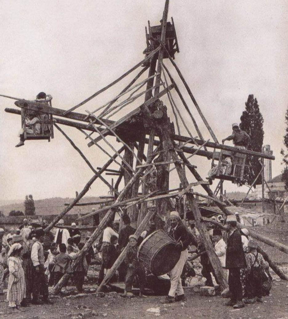 Historický drevený kolotoč s deťmi, obklopený skupinou ľudí počas zábavného jarmoku.