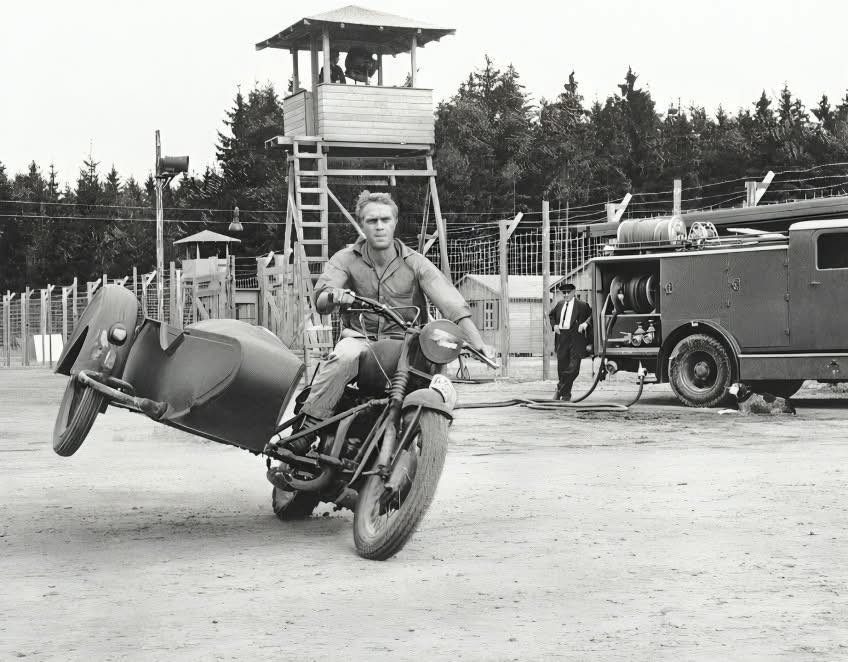 Muž na motorke so sajdkárou v pohybe, strážna veža a dodávka v pozadí. Retro scéna pripomínajúca filmový moment.
