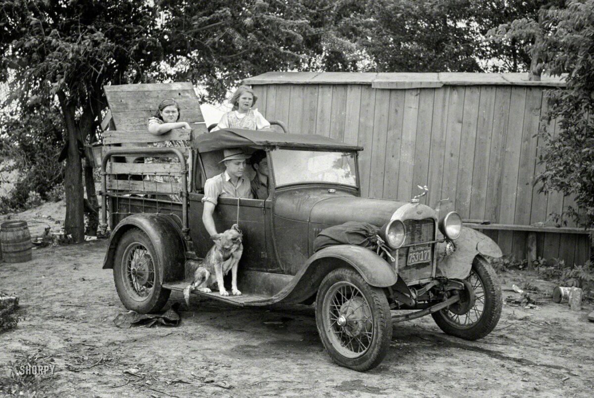 Staré auto s rodinou a psom, zaparkované pred drevenou ohradou, zachytené na čierno-bielej fotografii.