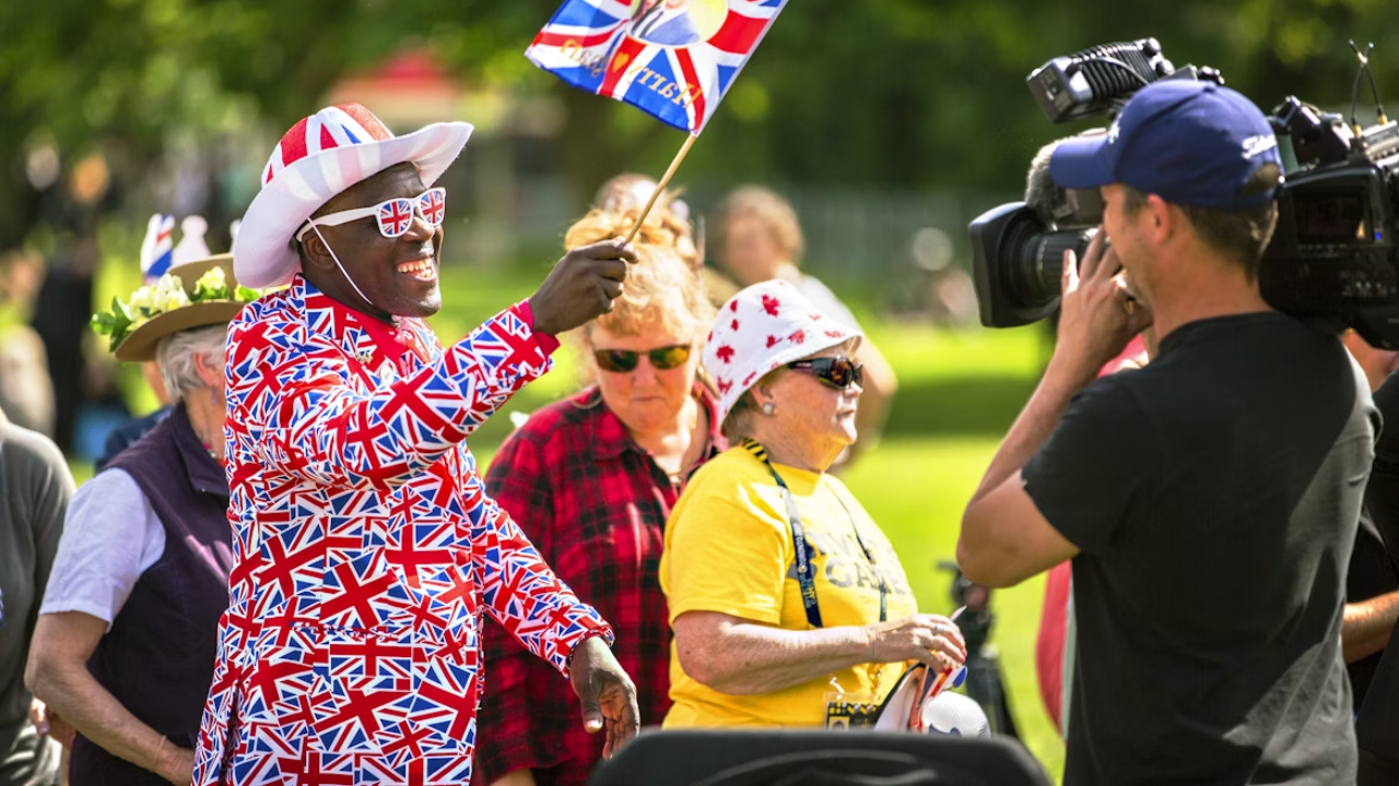 Osoba v oblečení s britskou vlajkou máva kameramanovi počas vonkajšieho podujatia.