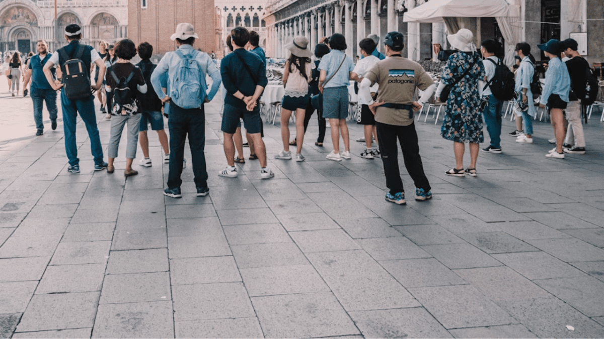 Skupina turistov na námestí v historickom meste, obdivujúci architektúru v letnom outfite.