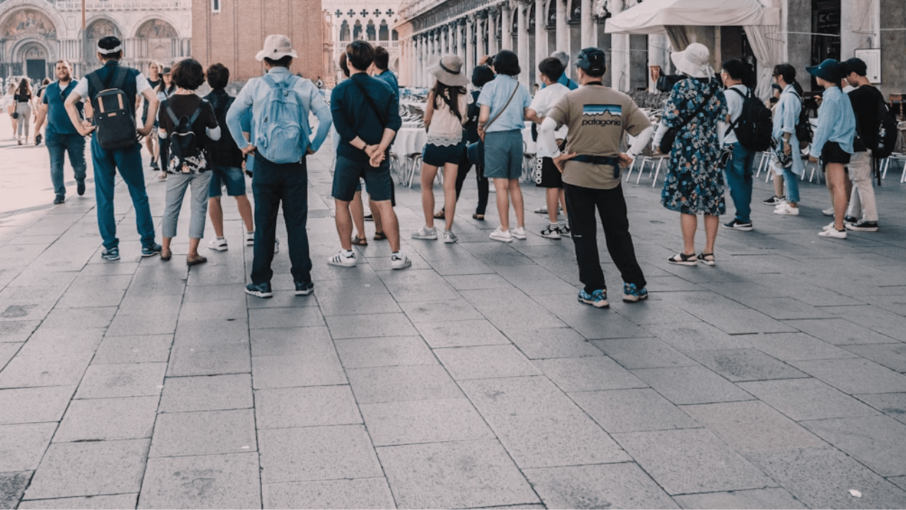 Skupina turistov na námestí v historickom meste, obdivujúci architektúru v letnom outfite.