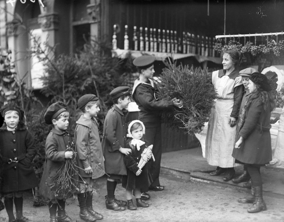Skupina detí pred obchodom s vianočnými stromčekmi, žena prijímá stromček od chlapca. Historická fotografia.