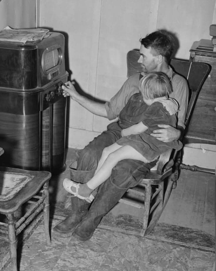 Muž s dieťaťom sedí v kresle a ladí starý rádio prijímač, vintage atmosféra v domácnosti.