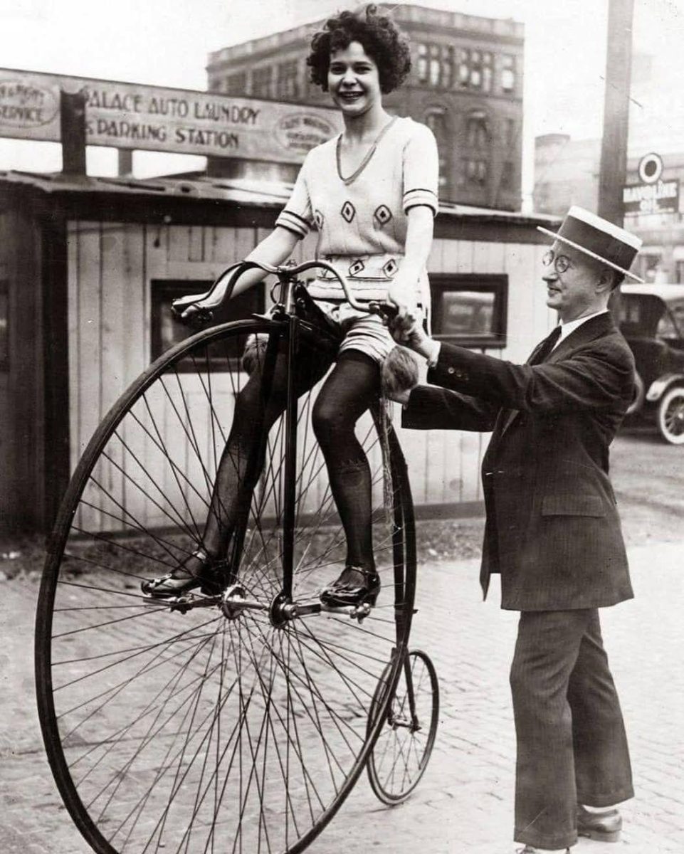 Žena na vysokých kolesách bicykla pomáhaná mužom, historická scéna z minulého storočia v mestskom prostredí.