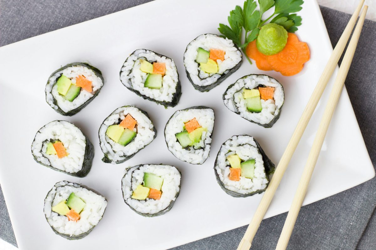 Vegánske sushi rolky s avokádom, mrkvou a uhorkou na bielom tanieri s lyžičkou wasabi a paličkami.