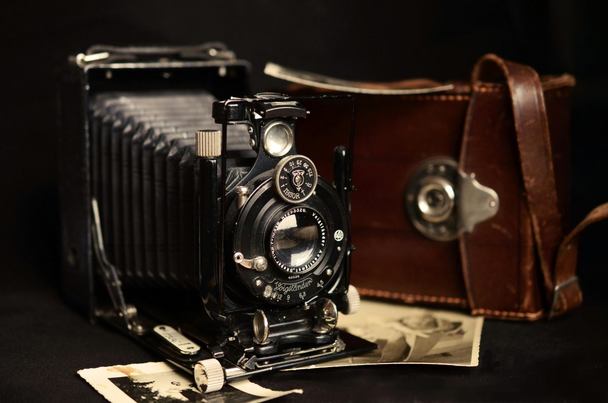 Stará analógová kamera a kožené puzdro na čiernom pozadí, pri ktorej sú položené staré fotografie.