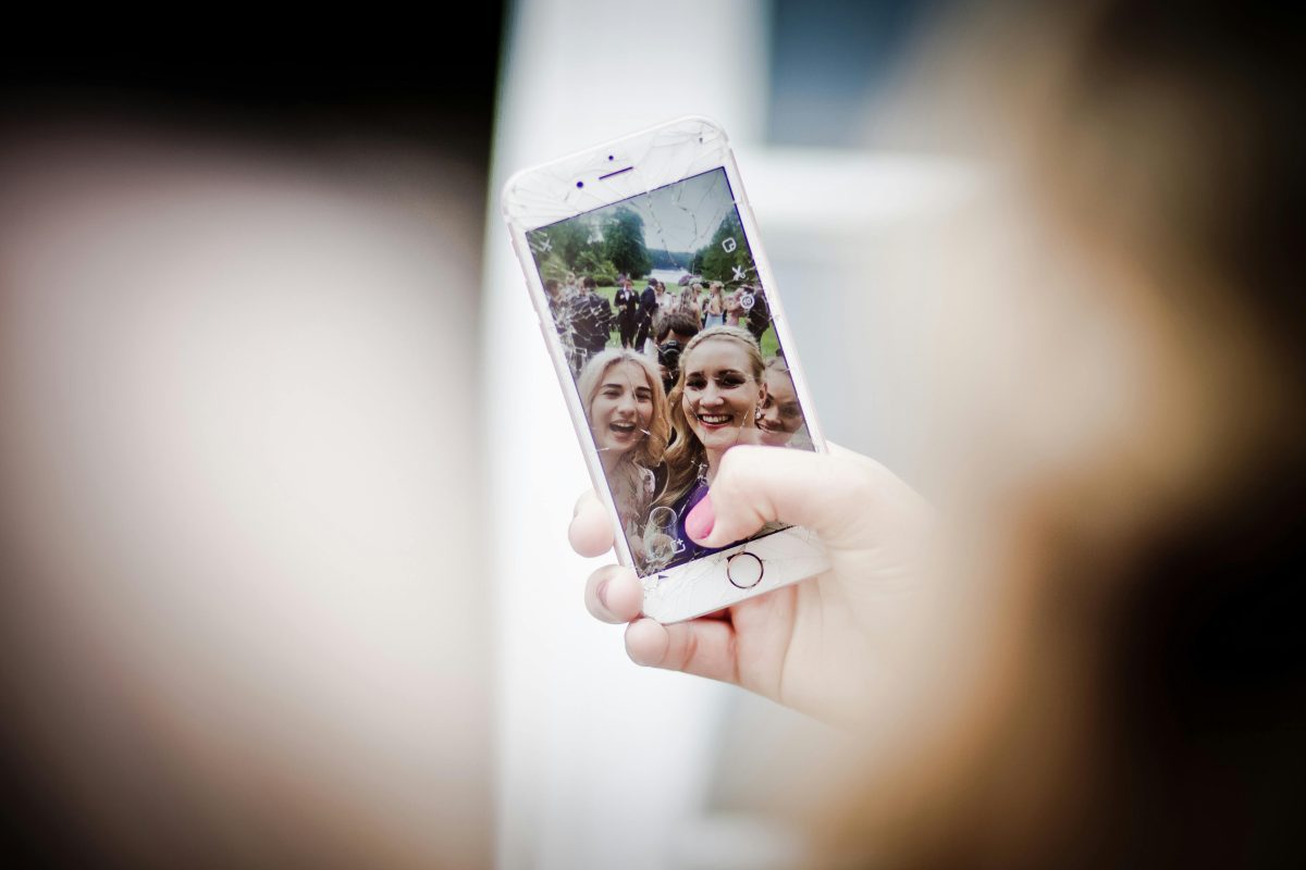 Žena si robí selfie vonku s priateľmi pomocou telefónu s prasknutým displejom.