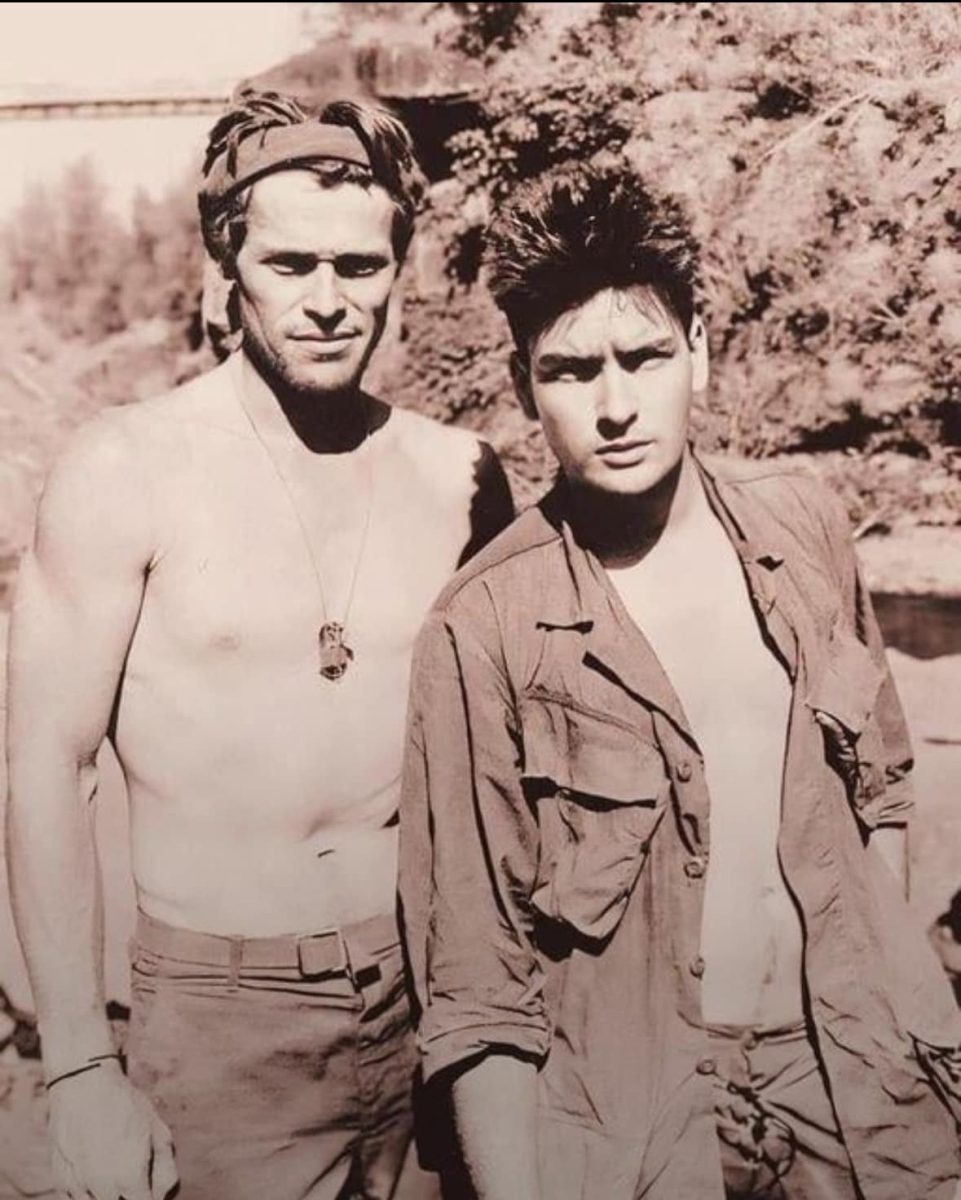 Dvaja muži v prírode pózujú pred kamerou, jeden je bez trička, druhý má košeľu. Retro štýl fotografie.