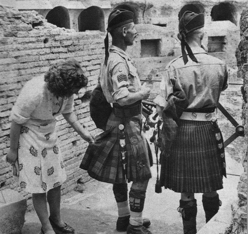 Žena skúma škótske kilty vo vojenskom prostredí, muži s tradičným krojom a nášivkami.
