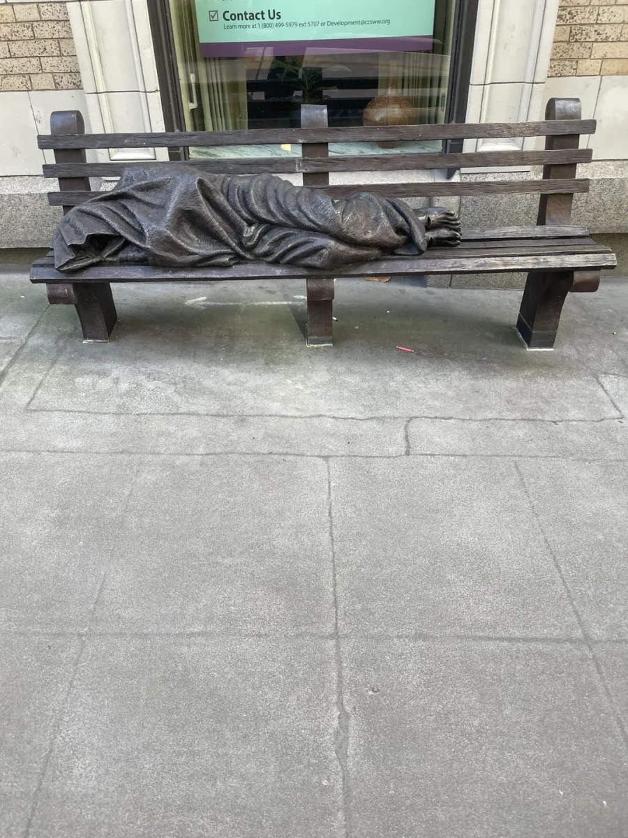 Socha prikrytého človeka ležiaceho na lavičke pred budovou, symbolizujúca bezdomovectvo.
